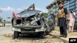 یو امنیتي سرتېری په باجوړ کې د بمي چاودنې له ښکار شوي ګاډي سره ولاړ دی - د جولای دویمې انځور.