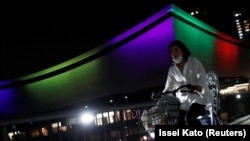 Женщина проезжает мимо Ариаке Арены, освещенной цветами олимпийских символов в день, знаменующий годичный обратный отсчет до Олимпийских и Паралимпийских игр в Токио 2020, которые были перенесены на 2021 год из-за вспышки коронавирусной болезни (COVID-19), 23 июля 2020 г. 