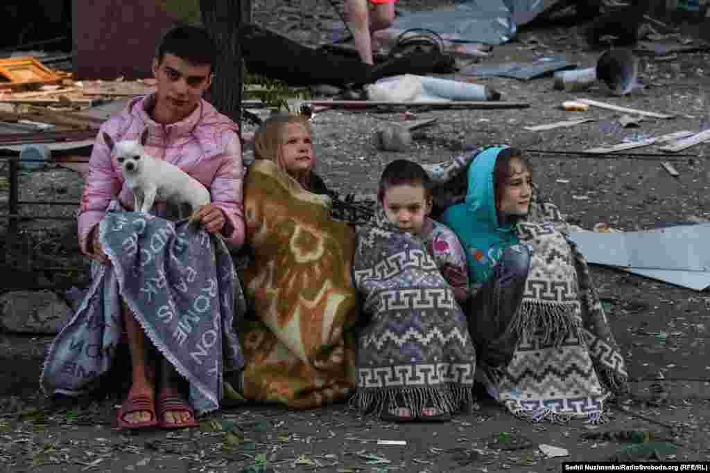 Çagalar hüjümde zeper ýeten jaýyň golaýynda ýorgana bürenip otyrlar. Olaryň öý iti hem ýanynda.