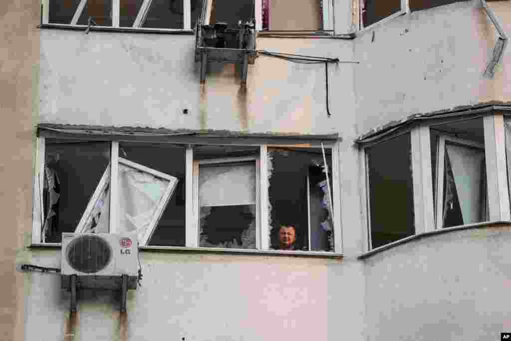مردی در حال نگاه کردن از پنجرهٔ یک ساختمان مسکونی چند طبقه که پس از حمله پهپادی روسیه به کی‌یف آسیب دیده است