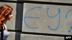 "ЕО?" деген жазудың қасынан өтіп бара жатқан әйел. Сурет Сербия астанасы Белград қаласында түсірілген.  