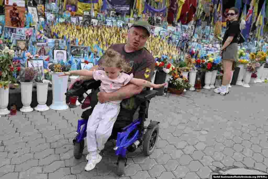 Вэтэран на вазку зь дзяўчынкай на руках побач з народным мэмарыялам на майдане Незалежнасьці ў Кіеве. Ліпень 2025 году